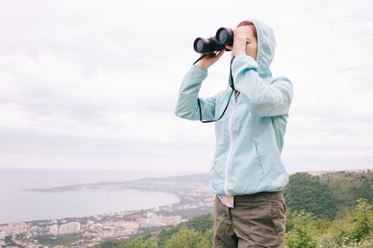 Traveler explorer young woman looking through binoculars on background of sea bay in the mountains. Copy-space in left part of image.