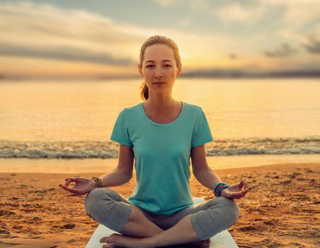 Young woman meditating in pose of lotus on yoga mat on sand beach near the sea at sunset in summer.