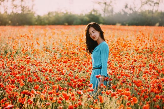 Summer beautiful brunette young woman walking through red poppy flower meadow, looking at camera.