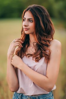 Portrait of beautiful sexy young woman with long brown hair in pink tank top and denim shorts posing outdoors at summer sunset, sensual, serious, blurry nature background