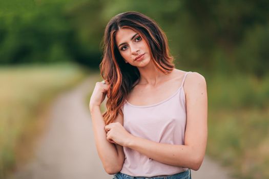 Portrait of beautiful sexy young woman with long brown hair in pink tank top and denim shorts posing outdoors at summer sunset, sensual, serious, blurry nature background