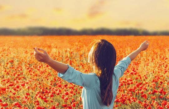 Happy young woman relaxing with raised arms in red poppies flowers meadow at sunset in spring.