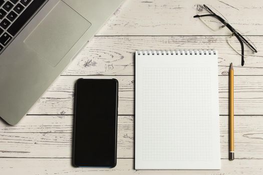Laptop computer with smartphone, eyeglasses, notepad and pencil on light wooden table, office stuff