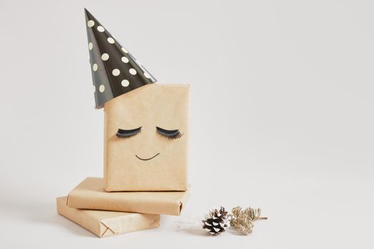 gift boxes with kraft paper on gray background, original gift decor with false eyelashes and party hat, minimalistic eco-friendly christmas decor, spruce branch and pine cone