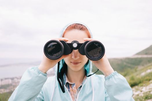 Explorer hiker young woman looking through binoculars on background of summer mountains outdoor, front view. Travel and journey concept.