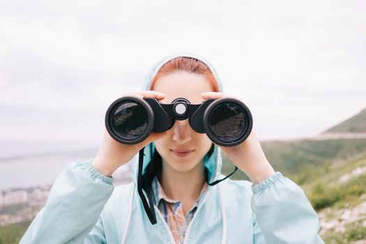 Explorer hiker young woman looking through binoculars on background of summer mountains outdoor, front view. Travel and journey concept.