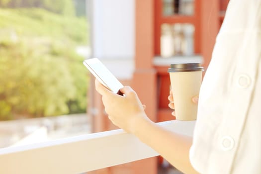 Unrecognizable woman using smartphone while standing with cup of coffee on balcony outdoor.