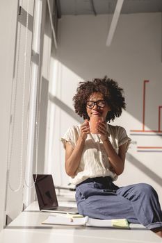 Happy Afro American woman holding cup of coffee while resting in the office during sunny day