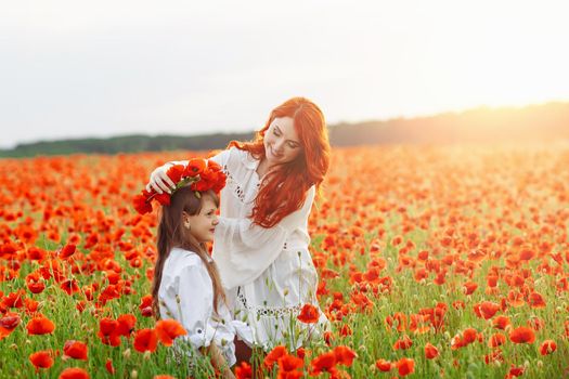 Little happy girl with redhead mother in white dresses makes wreath on poppy field at warm summer sunset