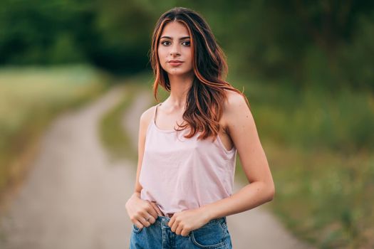 Portrait of beautiful sexy young woman with long brown hair in pink tank top and denim shorts posing outdoors at summer sunset, sensual, serious, blurry nature background