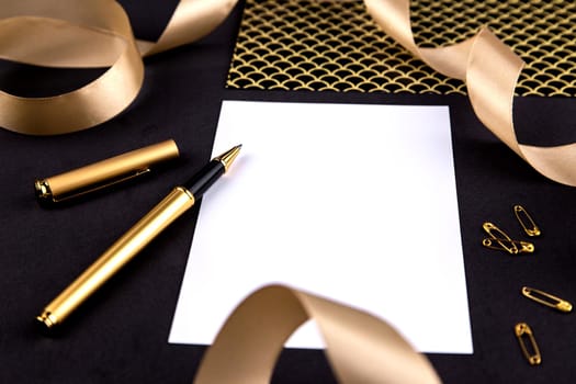 Gold pen, ribbon, paper clips and stationery on a black background with a white sheet of paper.