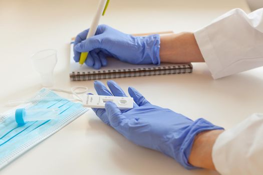 sars covid antigen test. mask and labglass at the table. doctor hands in blue gloves holding rapid test and writing results to note