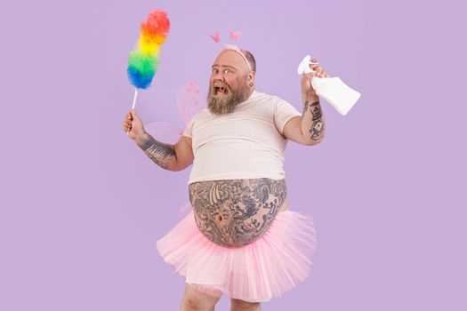 Emotional middle aged man with overweight in fairy costume with pink skirt holds pp duster and spray bottle on purple background in studio