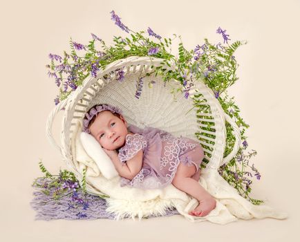 Adorable newborn baby girl wearing beautiful dress and wreath lying in basket with plant decoration holding hands under her cheeks in studio. Cute infant child with flowers