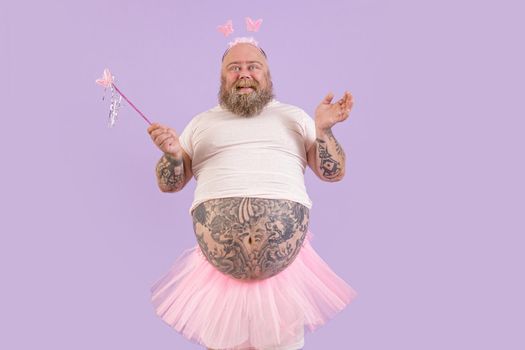 Smiling plus size mature man with tattooed abdomen wearing fairy suit holds magic stick with butterfly posing on purple background in studio
