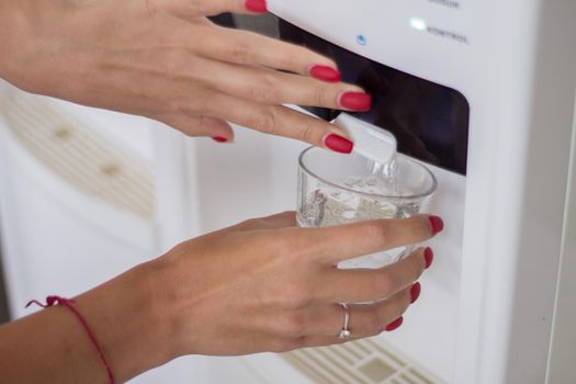 Female hand pours water from a water cooler in glass cup, close up