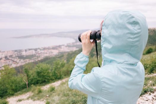 Unrecognizable traveler explorer young woman looking through binoculars on sea bay while walking in summer mountains.