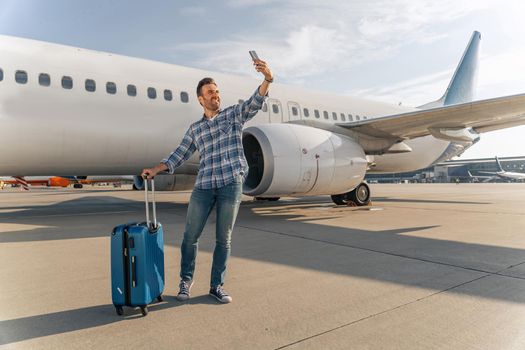 Smiling traveler standing with suitcase near plane while taking selfie in airport