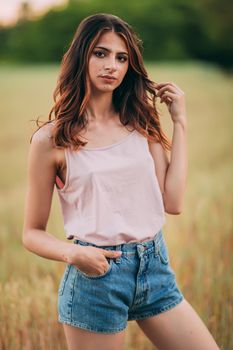 Portrait of beautiful sexy young woman with long brown hair in pink tank top and denim shorts posing outdoors at summer sunset, sensual, serious, blurry nature background