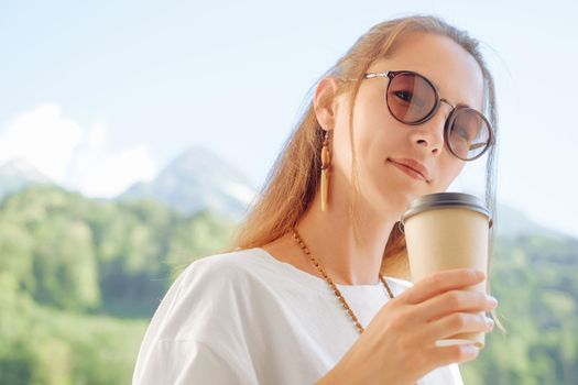 Beautiful young woman in sunglasses standing with cup of coffee on background of summer mountains, looking at camera.