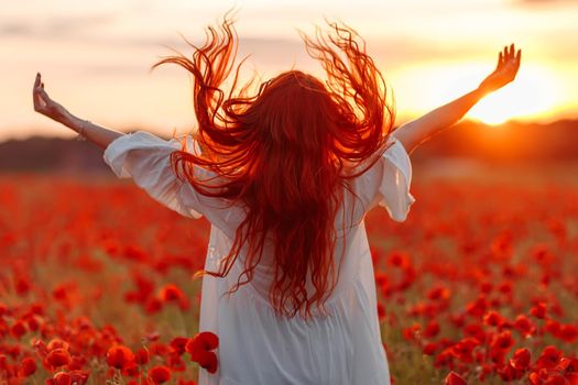 Happy redhead smiling woman in white dress on field of poppies at warm summer sunset