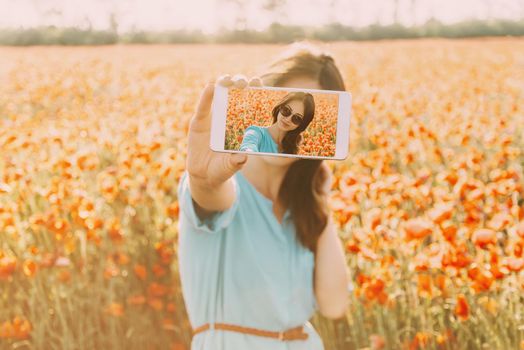 Beautiful brunette young woman in glasses taking a photo selfie with smartphone in flowers meadow in summer outdoor.