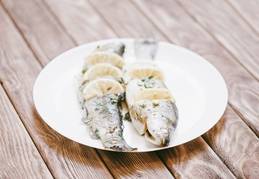 Fish trout dish with lemons and greenery on a white plate on wooden table.