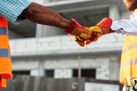 Two men engineers in workwear shaking hands against construction site, close up