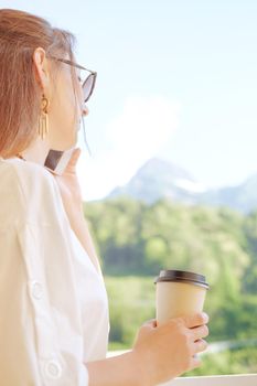 Young woman talking on phone and holding cup of coffee, mountains in the background.