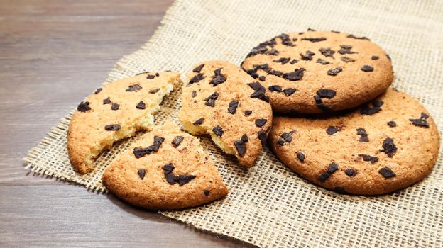American chocolate chip cookies on a brown wooden table and on a linen napkin close-up. Traditional rounded crunchy dough with chocolate chips. Bakery. Delicious dessert, pastries. Rural still life