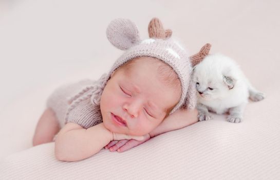 Adorable newborn baby boy sleeping on his tummy and little fluffy white kitten sitting close to him. Cute infant kid wearing knitted costume and hat napping with cat kity during studio photoshoot
