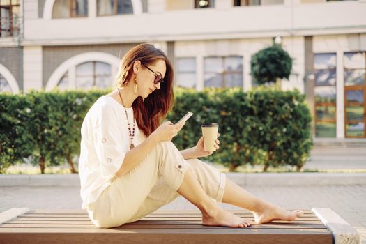 Beautiful brunette barefoot young woman sitting with cup of paper coffee on wooden bench and using mobile phone in summer, city lifestyle.