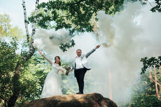 The bride and groom stand on a cliff and hold smoke bombs in their hands in white.