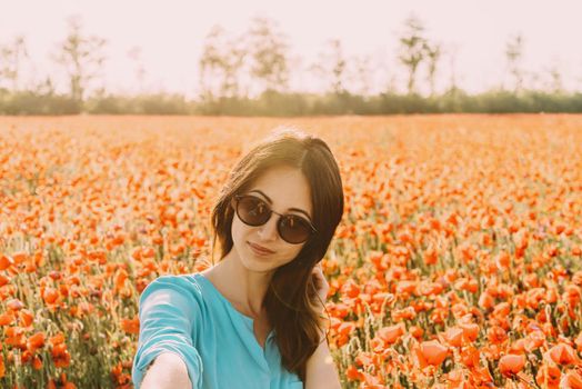 Beautiful smiling brunette young woman in glasses taking a self-portrait in flowers poppies meadow outdoor, point of view.