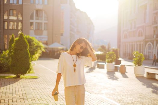 Happy beautiful young woman walking with paper cup of coffee in city in summer morning.