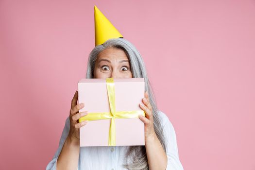 Happy middle aged Asian woman with grey hair and yellow party hat holds gift box near face on pink background in studio