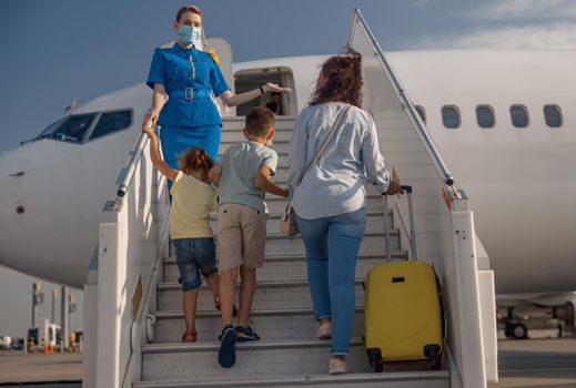 Back view of mother with two little kids and suitcase boarding the plane. Air stewardess wearing protective mask welcoming the family. People, traveling, vacation concept