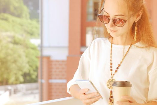 Beautiful boho stylish young woman using mobile phone while drinking coffee on balcony in spring morning, city lifestyle.