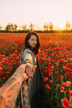 Beautiful brunette young woman holding male hand and leading him in poppy flower meadow at summer sunset, looking at camera. Point of view.