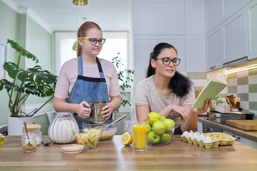 Mom and preteen daughter cooking together at home in kitchen apple pie, mother with book of recipes reading sequence of baking. Family, lifestyle, parent child relationship, homemade food concept