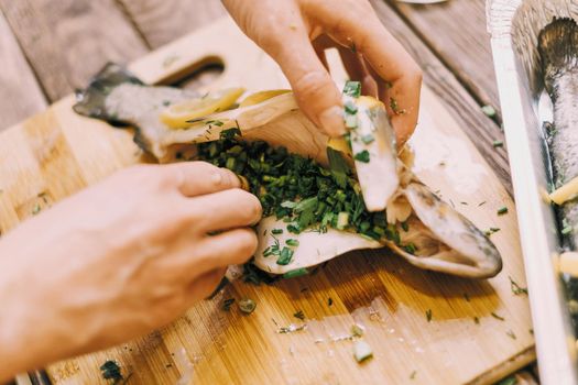 Unrecognizable woman putting greenery in raw fish trout for cooking in the kitchen, view of hands.