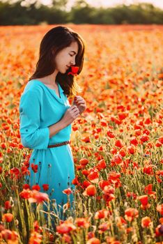 Dreamy smiling beautiful young woman smelling a red poppy in flower field in summer.