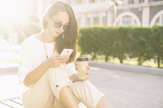Casual style girl using phone while sitting with paper cup of coffee in summer morning in city.