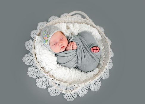 Adorable newborn baby girl wearing knitted hat lying on her back in basket and sleeping. Sweet swaddled in fabric infant child napping during studio photoshoot