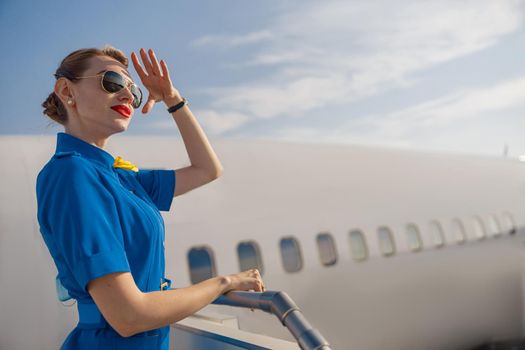 Portrait of elegant air stewardess in blue uniform and sunglasses holding hand near her head and looking far away, standing on airstair on a daytime. Aircrew, occupation concept