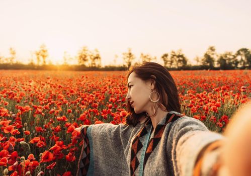 Point of view image of beautiful young woman making selfie in poppy flower field at sunset in summer outdoor.