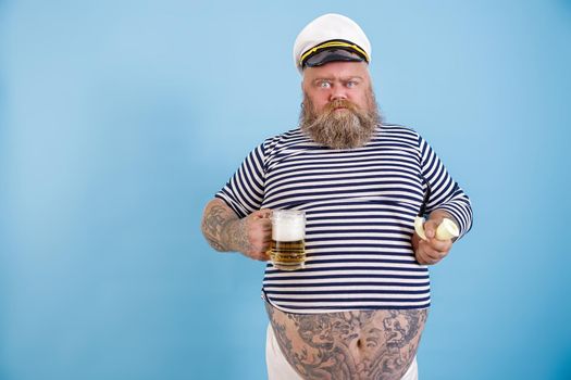 Funny obese man with beard in sailor costume and cap holds smoking pipe and glass of beer grimacing on light blue background in studio
