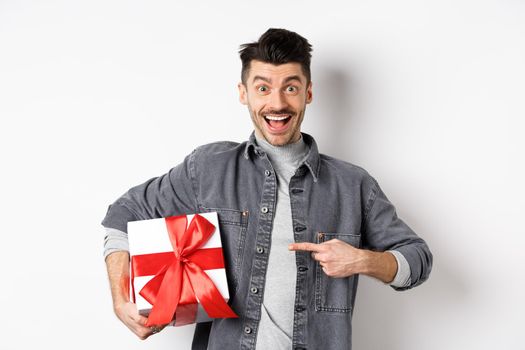 Happy Valentines day. Excited caucasian guy pointing at gift box from lover, smiling and looking amazed, buying presents on romantic date, white background.