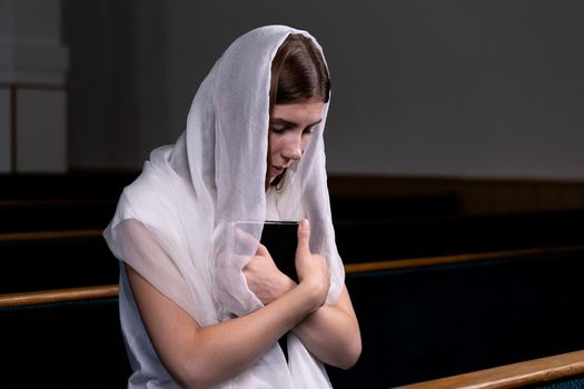 A young modest girl with a handkerchief on her head and a bible in her hands is sitting in church and praying. The concept of religion, prayer, worship.