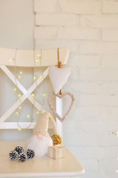 Cute gnome with long beard sitting on the chair near brick wall. white cristmas dwarf near hearts, pine cones and warm lights
